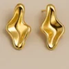 Pendientes Onda Dorada Par de pendientes stud esculturales de color dorado intenso con un diseño orgánico y fluido. La forma es asimétrica y abultada, similar a metal líquido o una gota, con un acabado pulido de alto brillo sobre un fondo beige claro.